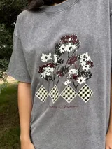 Remera de algodón gris con efecto lavado, de corte holgado y manga corta. Presenta un estampado frontal con diseño de árboles y figuras geométricas a cuadros, acompañado por la frase 'Jardín de Invierno'.