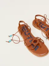 Sandalias planas de cuero color marrón, con diseño de punta cuadrada, tiras finas cruzadas y un detalle trenzado circular en el empeine. Cuentan con tiras para atar al tobillo decoradas con cuentas turquesas en las puntas.
