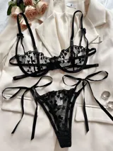 Conjunto de lencería negro de dos piezas con bordado de corazones y detalles de tiras con lazos. Incluye un sostén con aros y tirantes regulables, y una bombacha colaless ajustable con lazos en los laterales.