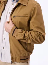 Campera tostada con cierre frontal, bolsillos laterales con cierre y un bolsillo adicional en el pecho. Cuenta con cuello clásico y puños y cintura elastizados con líneas oscuras contrastantes.