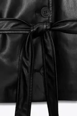 Chaqueta negra de efecto piel con cuello solapa, manga larga y trabillas en los hombros. Presenta bolsillos delanteros y cierre frontal con botones, ajustada a la cintura con un cinturón del mismo tejido.