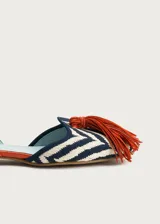 Sandalias planas destalonadas confeccionadas en rafia trenzada con patrón de zigzag en tonos azul marino y crudo. Tienen punta cerrada con una borla decorativa de flecos naranjas en el empeine, y una tira trasera ajustable de cuero naranja con hebilla metálica dorada.