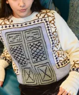 Sweater de lana merino tejido a mano, color marfil, con diseño jacquard geométrico en tonos marrón, violeta y gris en el pecho y las mangas. Cuenta con cuello redondo y mangas largas.