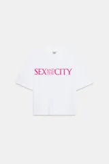 Camiseta blanca de manga corta y cuello redondo, con corte regular y bajo recto. Presenta el logo de "Sex & The City" estampado en color fucsia en el frente.