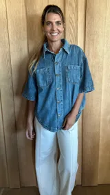 Sobrecamisa de denim soft en tono azul medio, con corte oversize, mangas cortas, cuello clásico y cierre frontal con botones metálicos. Presenta dos bolsillos frontales con solapa y un ruedo redondeado.