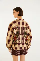 Chaqueta de cuello camisero, manga larga y cierre frontal oculto. Presenta un estampado de cuadros en tonos marrón y beige, complementado con un diseño bordado de flores y hojas en la parte delantera.