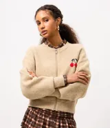 Cardigan de punto color beige con cierre frontal, cuello redondo, mangas largas y detalle bordado de cerezas en el pecho. Presenta puños y ruedo acanalados.