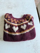 Sweater tejido a mano en mohair, de cuello redondo, con diseño de corazones y patrón de damero en la parte superior. Presenta una combinación de colores en tonos bordó, naranja y crema.