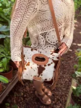 Bandolera de cuero con estampado animal print de vaca en tonos marrón y blanco, con flecos de cuero marrón en los laterales. Tiene correa larga de cuero marrón regulable y asa de mano con forma ovalada.