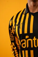 Camiseta de fútbol Puma del Club Atlético Peñarol, modelo Aniversario de manga larga. Diseño combinado con la mitad derecha a rayas verticales amarillas y negras, y la mitad izquierda totalmente negra con detalles en amarillo.