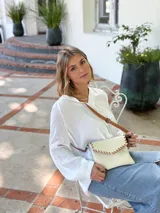 Cartera tipo sobre color crudo, confeccionada en cuero, con correa larga de cuero marrón y costuras decorativas en contraste.