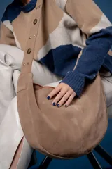 Bolso tipo bandolera de gamuza color marrón, con correa ancha ajustable decorada con tachas metálicas y cierre superior.