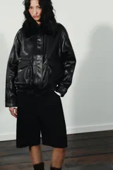 Cazadora de cuero sintético en color negro, con cuello de solapa y aplicación de pelo sintético desmontable. Presenta cierre frontal con cremallera y botones a presión ocultos, bolsillos de plastrón con solapa en el delantero y bajo con elástico.