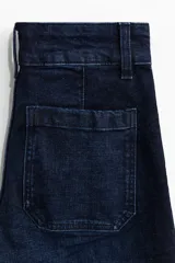 Pantalón de jean estilo wide leg con tiro alto, confeccionado en denim de algodón con leve stretch. Presenta cierre frontal con doble botón metálico, pliegues frontales y traseros, y bolsillos traseros de parche.