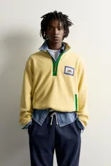 Sudadera polar de corte regular color amarillo, con cuello subido en tono azul, cierre frontal de cremallera verde y bolsillos con ribete verde. Incluye un parche gráfico de Fruit of the Loom en el pecho.