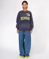Buzo de algodón y poliéster, color azul oscuro con efecto desgastado/tie-dye. Presenta un gran estampado frontal con la palabra "MICHIGAN" en amarillo con borde negro, un corazón naranja y apliques de pines decorativos en la parte superior. Tiene cuello redondo con terminación deshilachada y bolsillo canguro frontal.