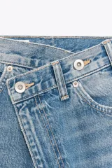 Jeans de tiro alto, color gris oscuro lavado, con pierna ancha extra larga (full length) y diseño de cintura cruzada asimétrica con cierre frontal de cremallera y botones metálicos.