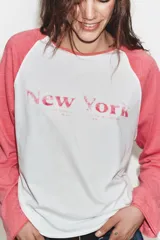 Camiseta de manga larga tipo ranglán con cuerpo blanco y mangas en color rosado. Presenta un estampado frontal con la inscripción 'New York' en tono rosado.
