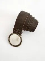 Cinto de cuero color chocolate oscuro con forro de cuero. Presenta una hebilla circular con borde metálico labrado y centro de guampa, con sistema de broche suizo intercambiable.