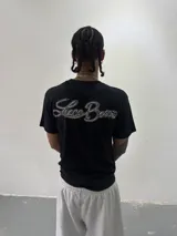 Remera de algodón color negro con cuello redondo y estampa gráfica blanca en el pecho. Presenta un corte regular y una pequeña etiqueta decorativa en la manga.