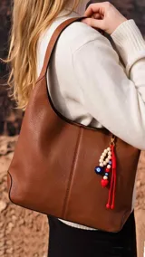 Cartera tipo hobo de cuero color marrón, con diseño de hombro, costuras visibles y llavero decorativo con cuentas de madera y dije de corazón rojo.
