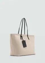 Bolso tipo shopper de gran tamaño confeccionado en tejido de canvas color beige. Cuenta con dos asas de hombro largas en contraste color negro, cierre de cremallera y un charm colgante decorativo en color negro.
