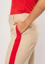 Pantalón beige de tiro alto con bolsillos delanteros y traseros, cierre frontal con cremallera y botón, y detalle de pespuntes rojos en los laterales.