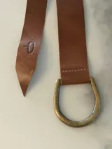 Cinto de cuero flexible y envolvente con diseño minimalista, que se anuda mediante una hebilla metálica en forma de D. El extremo presenta el logo de la marca grabado.