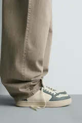 Zapatilla deportiva de estilo retro con diseño de paneles en combinación de colores crema, verde azulado y detalle en rojo en el talón. Presenta suela con volumen y cierre con cordones.