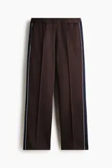 Pantalón deportivo marrón de punto con leve brillo, corte relajado y piernas rectas con pliegues cosidos. Cuenta con franjas laterales oscuras y ribetes blancos contrastantes, cintura elástica y bolsillos laterales.