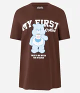 Remera de algodón color marrón con cuello redondo y manga corta. Presenta una estampa frontal de un osito de la serie Los Cariñositos sosteniendo una taza de café, acompañada por el texto 'My First Coffee' y 'Talk to me after ten o'clock'.