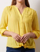 Blusa blanca de gasa marca Zac & Rachel, con cuello a la base, escote en V, abertura central con botones, bolsillo aplicado en el pecho y mangas largas regulables con presilla y botón. Tiene terminación opcional de nudo en el ruedo.