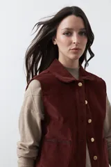 Chaleco de pana color marrón oscuro, con cuello camisero, cierre frontal de botones metálicos dorados y dos bolsillos frontales con detalle de bordado geométrico.