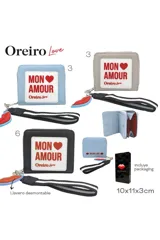 Billetera tipo monedero de cuero sintético negro con parche frontal blanco que incluye el texto 'MON AMOUR' y logo de la marca. Incluye una correa de muñeca a rayas y un dije colgante en forma de labios rojos.