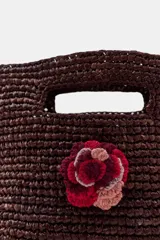 Mini bolso de mano tejido en rafia color marrón oscuro, con diseño estructurado y detalle de flor decorativa en tonos rojizos realizada en crochet.