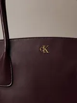 Cartera tote Calvin Klein de color marrón oscuro, con doble asa de hombro, cierre de cremallera superior y monograma metálico dorado en la parte frontal.