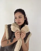 Chaleco estilo gamulán de cuero sintético marrón con interior y bordes de corderito sintético color crema. Presenta cierre frontal con botones a presión y bolsillos laterales.