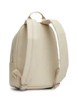 Mochila Tommy Jeans de diseño minimalista en color beige, confeccionada en poliuretano de acabado liso. Cuenta con un compartimento principal con cierre, un bolsillo frontal con cremallera, asa superior y correas ajustables. Incluye el logo metálico de la marca en la parte inferior frontal.
