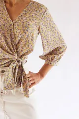 Blusa cruzada con estampado floral en tonos de lila, amarillo y verde. El cruce se ajusta a la cintura con un lazo. Tiene mangas 3/4 con puños elásticos y escote en V.