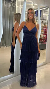 Vestido largo de fiesta color azul marino, con acabado brillante y textura plisada. Presenta escote en V, breteles finos y falda con tres niveles de volados.