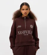 Buzo de algodón y poliéster color marrón, con cuello alto y cierre frontal parcial. Presenta diseño de hombros caídos, puños y ruedo acanalados, y estampa gráfica frontal con lettering Nashville.