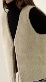 Chaleco sin mangas de corderito sintético color beige, con frente de textura suave y espalda de gamuza. Diseño abierto sin cierres.
