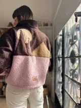 Buzo de tejido tipo corderito con diseño de bloques de color en tonos marrón, chocolate y rosa. Presenta un aplique decorativo en la espalda con forma de paisaje montañoso y etiqueta de marca en el ruedo.
