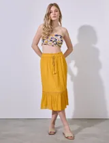 Falda midi color mostaza, de gasa bambula con pretina smock, lazo de ajuste, volado en el ruedo y aplique de puntilla.