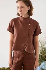 Blusa de lino color marrón, de corte recto y estilo japonés, con mangas cortas, cuello alto con volados y cierre lateral cruzado con pequeños botones dorados. Presenta costuras a la vista en contraste.