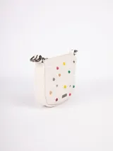 Cartera bandolera color crema de símil cuero, con apliques frontales de tachas redondas de colores y estrellas plateadas. Tiene correa regulable con hebillas y cierre superior con cremallera.