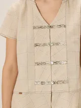 Camisa de manga corta color beige con estampado a cuadros, cuello en V y detalles de galones decorativos estilo oriental en la parte frontal.