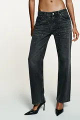 Jeans de tiro bajo y corte recto, confeccionados en denim color gris oscuro con efecto lavado. Presentan cinco bolsillos y están decorados con aplicaciones de strass multicolor en diferentes formas y tamaños en la parte frontal. Cierre frontal con cremallera y botón metálico.