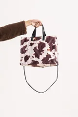 Cartera tipo tote con estampado de vaca, confeccionada en material con textura similar al pelo. Cuenta con asas cortas fijas de color negro y una correa larga desmontable y ajustable, también en color negro.