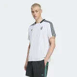 Remera blanca con finas rayas verticales celestes, cuello redondo negro con ribete verde y mangas cortas con ribete negro. Presenta las tres tiras negras características de Adidas en los hombros, el logo de Adidas bordado en el pecho y el escudo del Liverpool FC bordado en el pecho.